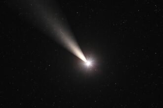 A high-resolution image of Comet 3I/ATLAS with a symmetric jet structure, set against a starry night sky with the Milky Way galaxy visible in the background.