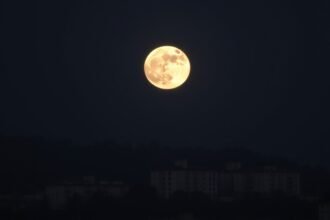 A high-quality image of the Wolf Moon supermoon rising over a cityscape or a natural landscape, with a clear view of the full moon and its surrounding environment.