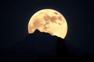 A high-resolution image of the full moon rising over a mountain range, with a subtle silhouette of a wolf in the foreground, symbolizing the Wolf Supermoon.
