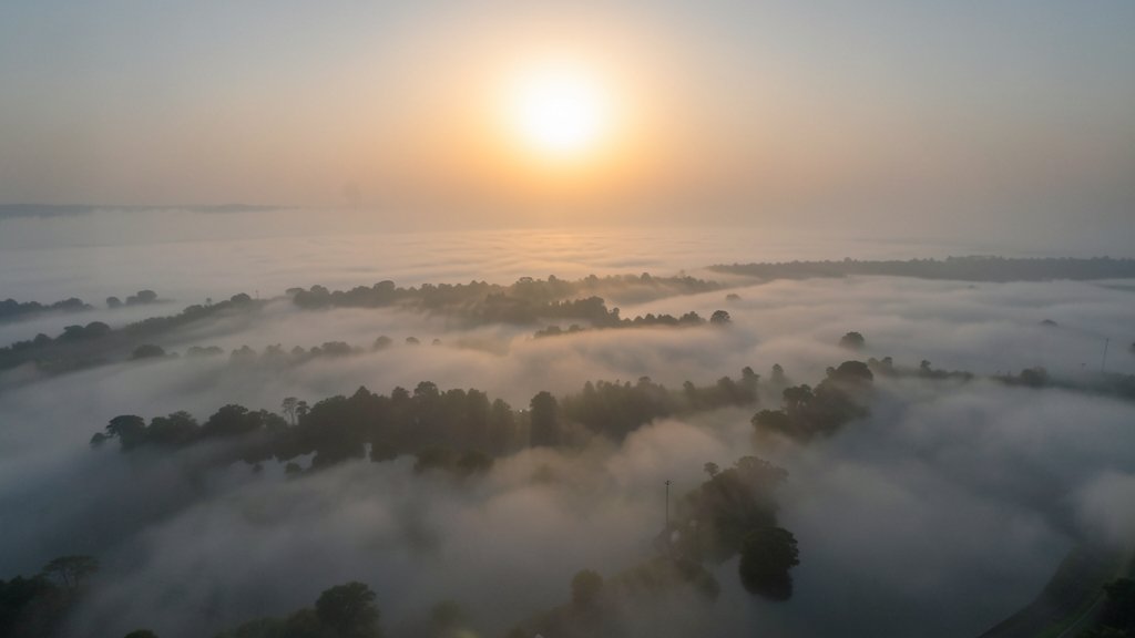 A satellite image of the Ganges Delta covered in a thick layer of fog, with the sun rising in the background, highlighting the contrast between the natural beauty and the environmental challenges faced by the region.