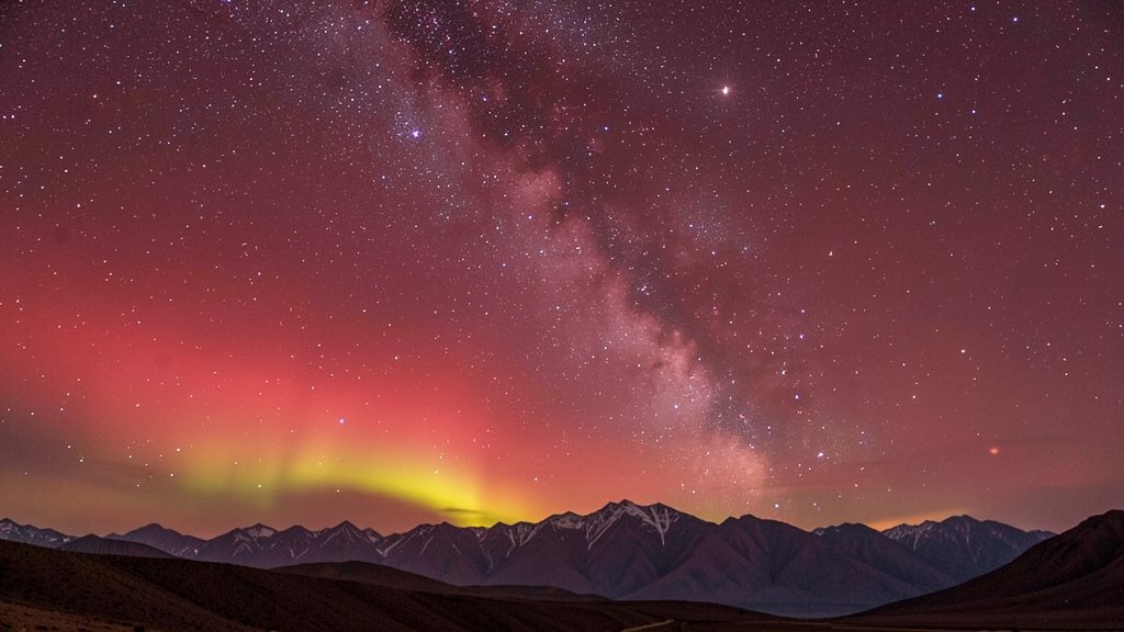 A photograph of the red sky over Ladakh's Hanle, with the Milky Way visible, and a few stars twinkling, during an auroral display, with a subtle hint of the Himalayan mountains in the background.